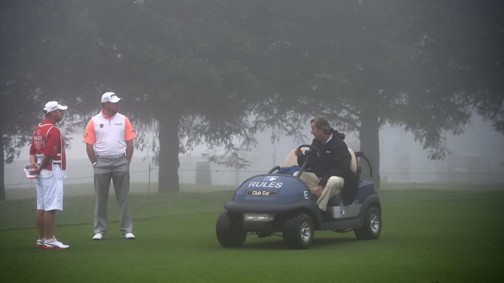 Lee Westwood of England and caddie Billy Foster talk with a rules official as they wait in the fog during the first round in Crans